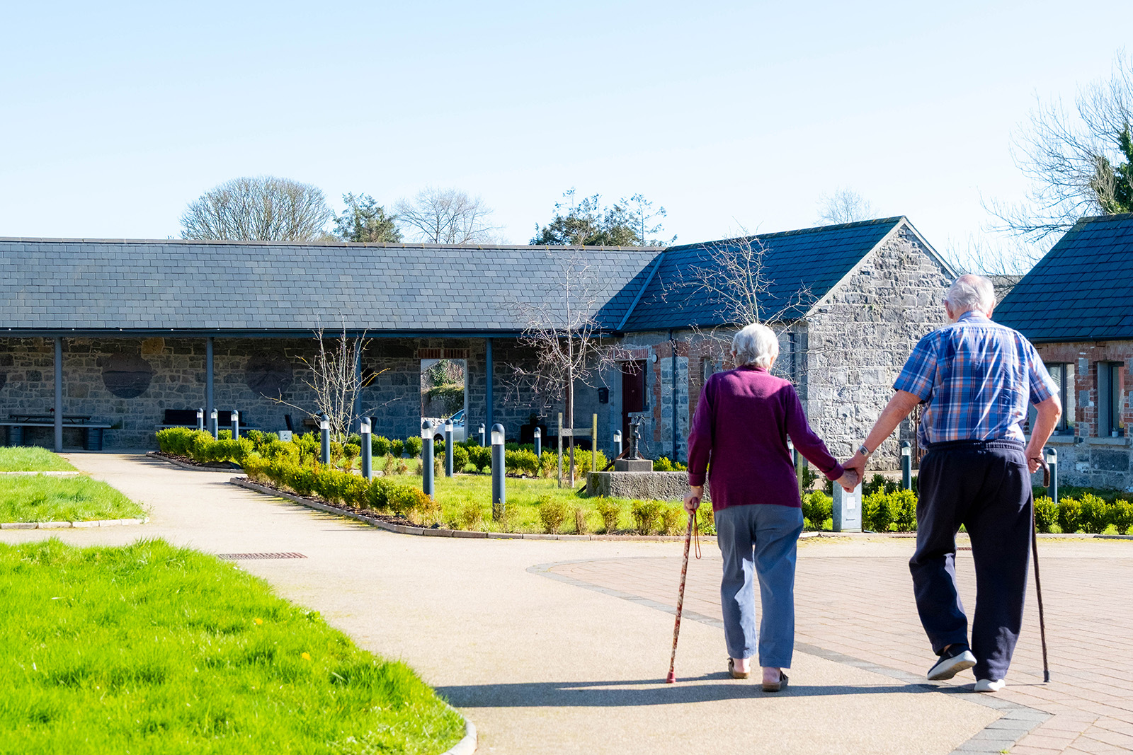 Wide shot of elderly man and woman from the back holding hands with Cuan An Chlair Centre buildings in background.